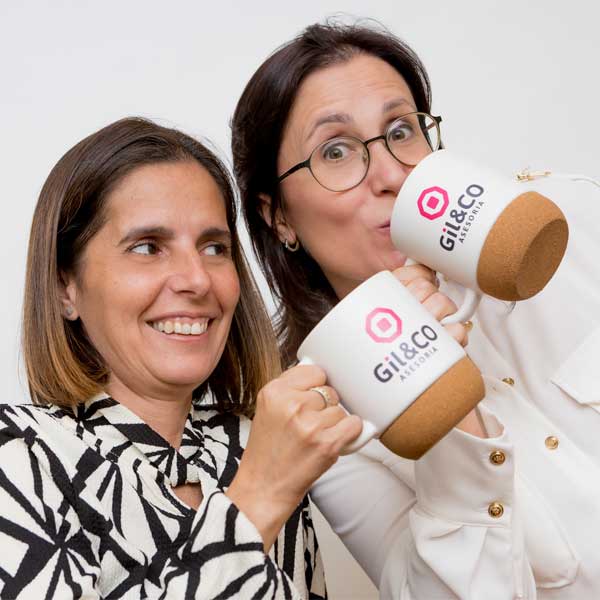 Dos mujeres sonríen y sostienen tazas blancas con la marca "Gil & Co" sobre un fondo liso.